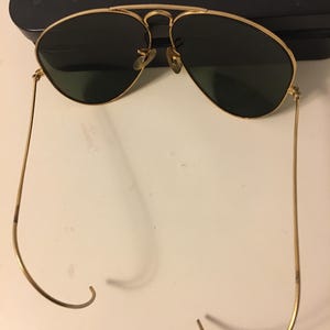 May include: Gold-framed aviator sunglasses with dark green lenses. The sunglasses have a double bridge and curved temples. The lenses are teardrop-shaped. The frame is a shiny gold color.