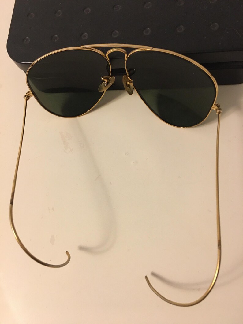 May include: Gold-framed aviator sunglasses with dark green lenses. The sunglasses have a double bridge and curved temples. The lenses are teardrop-shaped. The frame is a shiny gold color.