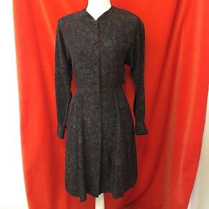 May include: A long-sleeved dress in a dark brown hue featuring a repeating floral pattern in shades of teal and muted green. The garment includes a button-down front closure, a simple collarless neckline, and long sleeves with buttoned cuffs.