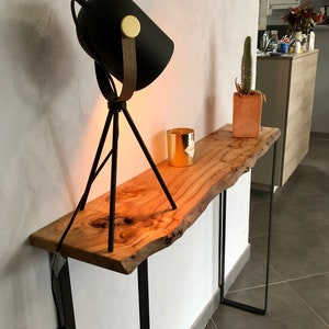 Peut inclure: Une console en bois avec un plateau en bois brut et des pieds en métal noir. Une lampe noire à trépied avec un accent doré est posée sur la table, ainsi qu'un bougeoir doré et un cactus en pot.