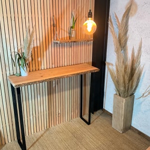 Peut inclure: Une console en bois avec un plateau en bois brut et des pieds en métal noir. La console est placée contre un mur en lattes de bois avec une petite étagère au-dessus. Une ampoule pend du plafond au-dessus de la table.