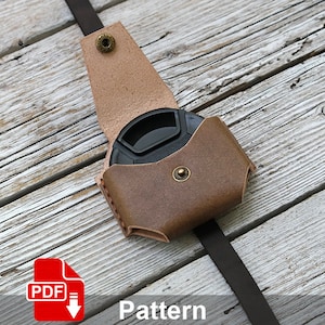 May include: A brown leather camera lens pouch with a flap and a snap closure. The pouch is lying on a wooden surface with a black strap attached.