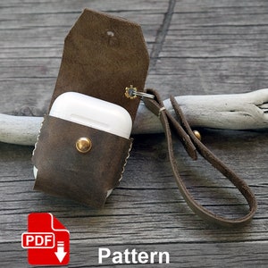 May include: A brown leather case with a snap closure for an AirPod case. The case has a leather strap with a metal ring. The case is sitting on a wooden surface. The text "PDF Pattern" is visible below the case.