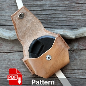 May include: A tan leather camera lens cap holder with a snap closure. The holder is open, revealing a black lens cap inside. A white strap is attached to the holder. The word "Pattern" is printed in white text.