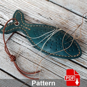 May include: A teal leather fish-shaped pincushion with white stitching and brown leather cord. The pincushion is filled with needles and thread. The image includes the text "Pattern" and a red PDF download icon.