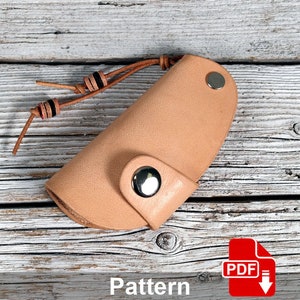Könnte beinhalten: Ein hellbrauner Leder-Schlüsselanhänger mit Druckknopfverschluss und einem hellbraunen Lederband mit schwarzen Perlen. Der Schlüsselanhänger liegt auf einer hellbraunen Holzoberfläche. Muster PDF.