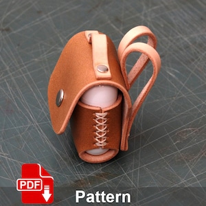 May include: A brown leather case with a stitched design and a strap. The case is designed to hold a small cylindrical object, such as a phone charger or a small flashlight. The case has a metal snap closure and a loop for attaching a carabiner.