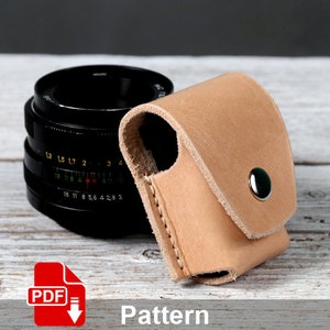 Puede incluir: Una funda de cuero marrón claro para objetivo de cámara con cierre de presión. La funda está sobre una superficie de madera blanca junto a un objetivo de cámara negro.
