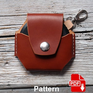 May include: A brown leather case with a snap closure and a metal loop for a key chain. The case is designed to hold a small round object, such as a coin or a button. The case is sitting on a wooden surface. Pattern.