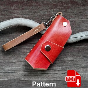 May include: A red leather key fob with a brown leather strap and a silver snap closure. The key fob is on a wooden surface. Pattern.