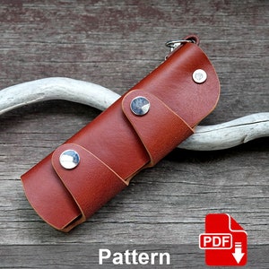 May include: A brown leather key holder with a silver snap closure. The key holder is folded and has a silver key ring attached. The key holder is on a wooden surface.