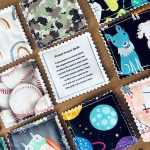 May include: A handmade quilt with different fabric squares. Each square has a different design, including a baseball, a rainbow, a camouflage pattern, a dog, and a space scene. The quilt has a pocket with a note that reads "Pocket Prayer Quilt I put prayers in every stitch the angels blessed each one so place the quilt in your hand and let God's presence come. Guardian angels gather close as they have a job to do so if you are scared or lonely this quilt can comfort you."