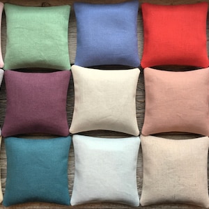 May include: A collection of 12 square-shaped pillows in various colors, including light pink, green, blue, red, beige, and gray. The pillows are made of a soft, textured fabric and are arranged in a grid pattern.