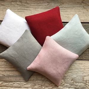 May include: Five square-shaped linen pillows in different colors: white, gray, red, light green, and pink. The pillows are arranged in a circle on a wooden surface.