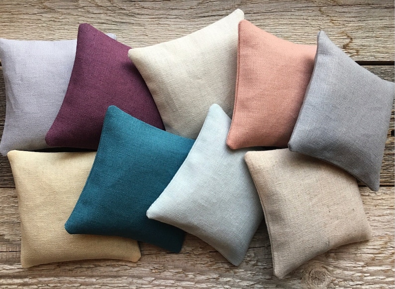 May include: Eight square throw pillows in various colors and textures, including beige, light gray, teal, light blue, burgundy, and peach. The pillows are arranged in a semi-circle on a wooden surface.