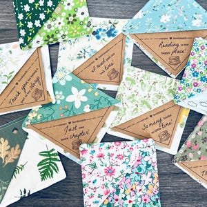 May include: A collection of colorful corner bookmarks with floral and botanical patterns. Each bookmark features a brown paper triangle with a printed quote about reading, such as "Thank you for being part of my story" and "Reading is my happy place!"