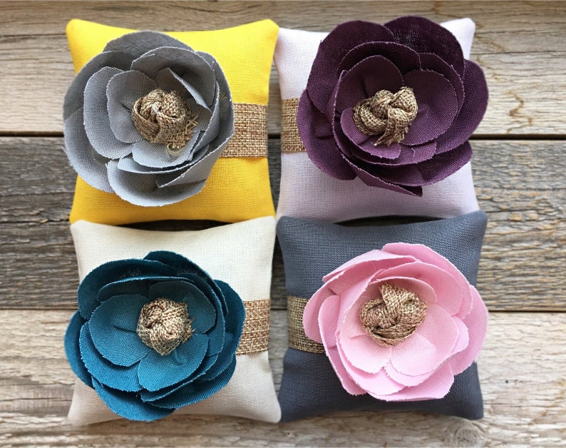 May include: Four small square pillows with fabric flower embellishments. The pillows are in shades of yellow, purple, beige, and gray. Each pillow has a burlap ribbon tied around the center.