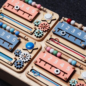 May include: A wooden activity board with four different sections, each featuring a different activity. The top left section has a zipper, a bead string, and a number line with the numbers 1 through 5. The top right section has a star-shaped button, a set of gears, and a number line with the numbers 1 through 5. The bottom left section has a star-shaped button, a set of gears, and a number line with the numbers 1 through 5. The bottom right section has a star-shaped button, a set of gears, and a number line with the numbers 1 through 5.