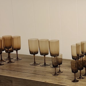 May include: A collection of brown-tinted glassware, including wine glasses and smaller stemmed glasses, arranged on a weathered wooden surface. The glasses have a simple, elegant design, with a slightly flared rim and a dark brown base. The background is a plain white wall.