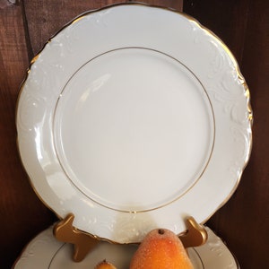 May include: A stack of white ceramic dinner plates with gold trim. The top plate has a raised floral design. Two pears, one yellow and one orange, are sitting on the top plate.