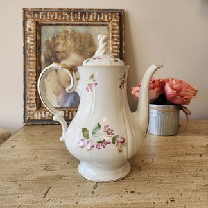 May include: White porcelain teapot with a gold trim and floral design. The teapot has a lid with a knob and a spout. The teapot is sitting on a wooden table.