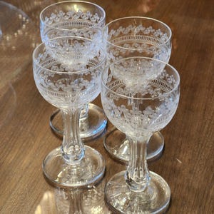 May include: Four clear glass cordial glasses with etched floral and scroll designs. The glasses have a round base, a slender stem, and a bowl-shaped top. The glasses are arranged on a wooden surface.