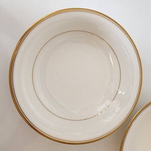 May include: Two white ceramic bowls with a gold rim and a gold line around the center. The bowls are empty.