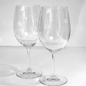 May include: Two clear glass wine glasses with a tall, thin stem and a wide bowl. The glasses are empty and sitting on a white surface.