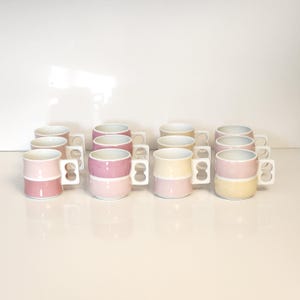 May include: A set of ceramic mugs in shades of pink and yellow. Each mug has a two-tone design and a white handle. The mugs are arranged in rows, highlighting their colour combinations and modern design. The mugs are suitable for tea or coffee.