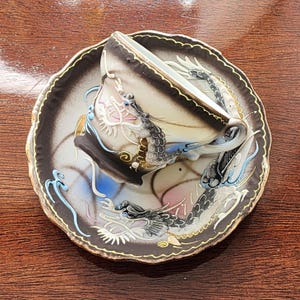 May include: A white teacup and saucer with a black and gold dragon design. The cup and saucer are decorated with a swirling pattern of blue, pink, and gold.