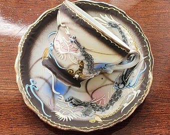 May include: A white teacup and saucer with a black and gold dragon design. The cup and saucer are decorated with a swirling pattern of blue, pink, and gold.