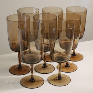 May include: A set of eight tall drinking glasses made of translucent brown glass. Each glass features a cylindrical bowl, a slender stem, and a wide circular base. The glasses are arranged in a cluster, showcasing the uniform amber-toned glass.