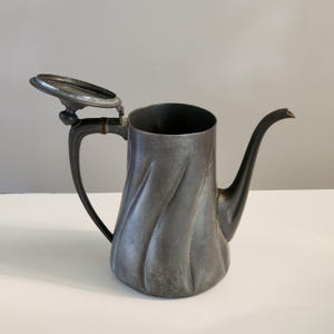 May include: A metallic gray pewter pitcher featuring a spiraling fluted body design, a curved handle, a long slender spout, and an attached hinged lid that is currently open. The surface has a matte, weathered finish with visible dark patina.