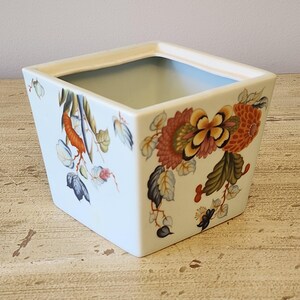 May include: A white square ceramic planter with a floral design. The design features orange, yellow, green, and blue flowers and leaves.