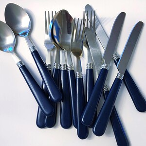 May include: A collection of stainless steel flatware featuring dark navy blue plastic handles with silver-toned ribbed accents at the neck. The set includes multiple spoons, forks, and serrated knives arranged in a scattered, overlapping pile.
