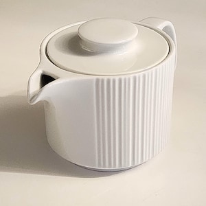May include: A white ceramic teapot featuring a cylindrical body with vertical ribbed detailing, a matching circular lid with a central knob, and a small integrated spout. The teapot has a smooth, glossy finish and a simple, functional design.