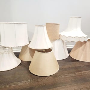 May include: A collection of various lamp shades in neutral tones. The shades vary in shape, including drum, bell, and empire styles. Some shades are white, others are beige or tan, and some have decorative trim.