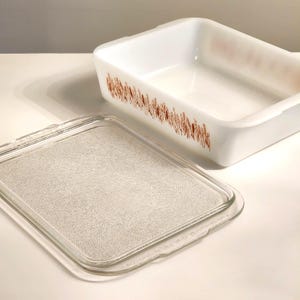 May include: A rectangular white milk glass baking dish with a brown wheat stalk pattern along the exterior sides. The dish includes a clear, textured glass lid with a smooth border, designed to fit securely over the top of the baking vessel.