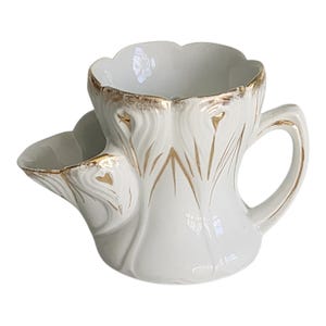 May include: White ceramic shaving mug with a unique design. The mug features a built-in soap dish and a handle. Gold accents adorn the rim and the decorative floral pattern. The mug is a vintage item.