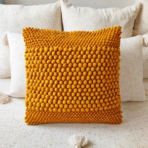 May include: A mustard yellow throw pillow with a textured surface made of small, round balls. The pillow is sitting on a white couch.