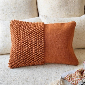 Puede incluir: Una almohada de sofá naranja quemado con una superficie texturizada y abultada en un lado y una superficie lisa y tejida en el otro. La almohada está sentada en un sofá blanco.