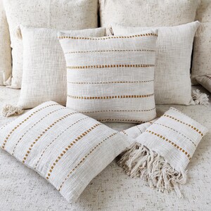 May include: A set of four throw pillows with a white background and gold stripes. The pillows are made of a soft, textured fabric and have a fringe trim.