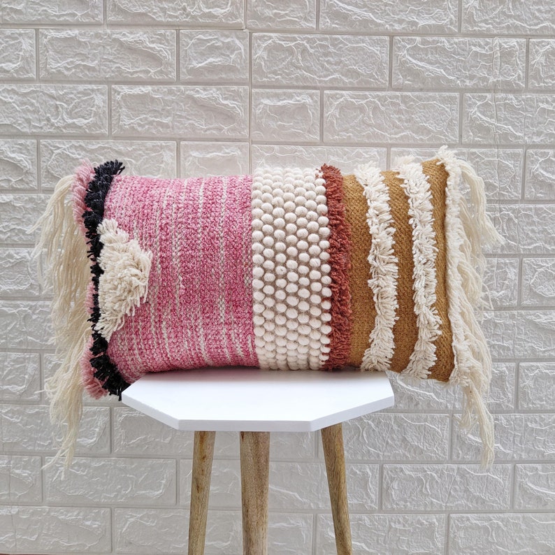 May include: A pink, white, and brown woven throw pillow with fringe detailing. The pillow is sitting on a white hexagonal table with wooden legs.