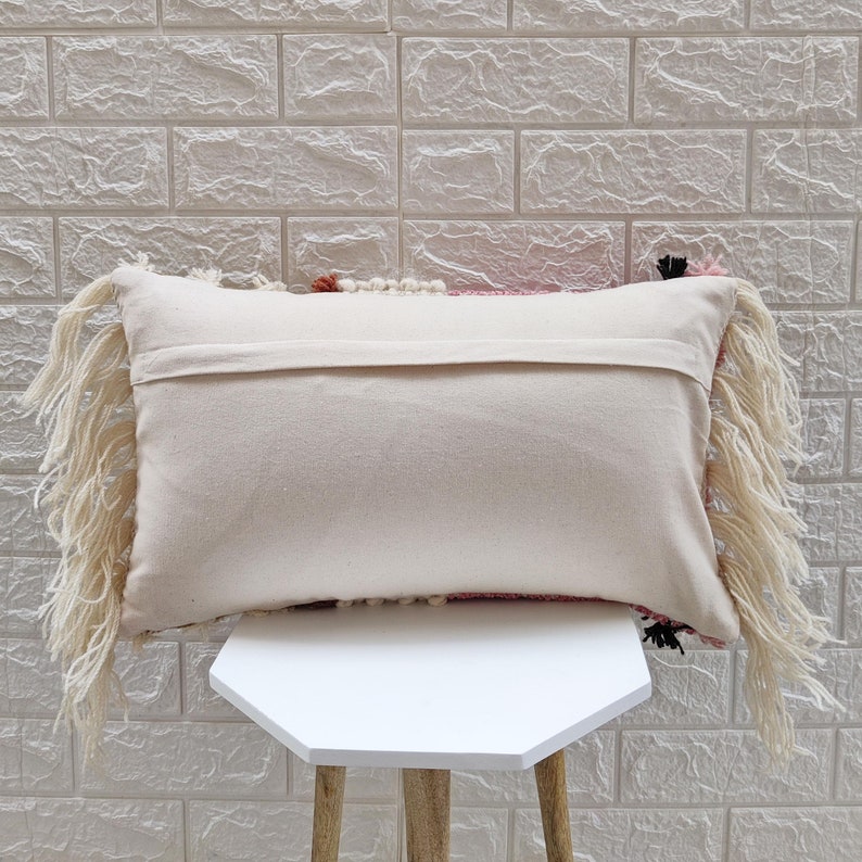 May include: A pink, white, and black throw pillow with fringe detailing. The pillow is sitting on a white stool.