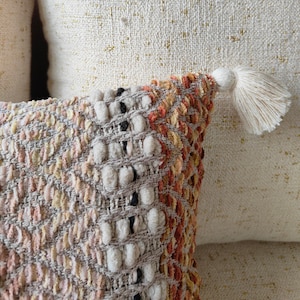 May include: A beige and brown throw pillow with a textured woven design and a fringe tassel. The pillow has a subtle ombre effect with a lighter beige colour at the top and a darker brown colour at the bottom.