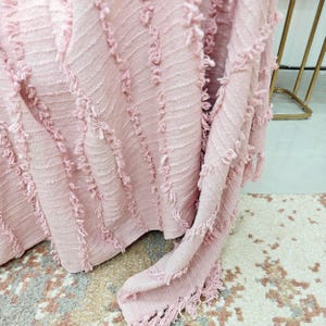 Puede incluir: Una manta tejida rosa pálido con un diseño texturizado y detalles de flecos. La manta está drapeada, mostrando su textura tejida y los flecos en los bordes. El color es un rosa suave y empolvado, ideal para una cama o sofá.