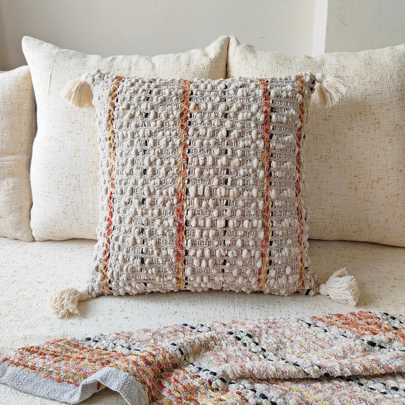 May include: A cream-coloured throw pillow with a textured woven design featuring orange and brown stripes. The pillow has tassels on each corner.