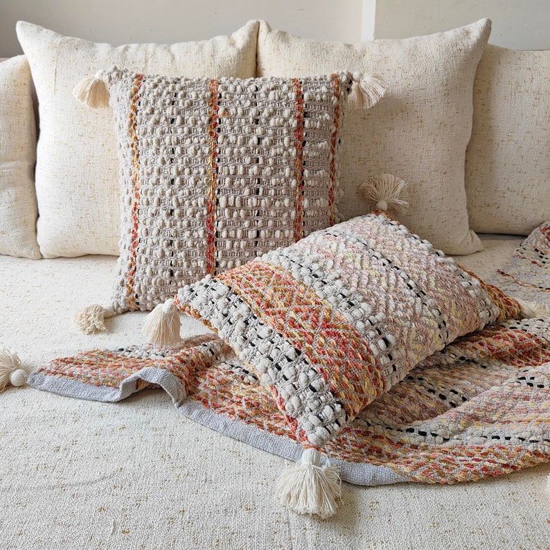May include: Three throw pillows with a woven texture and fringe tassels. The pillows are on a white sofa with a cream-colored throw blanket.