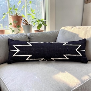 May include: A long, rectangular black pillow with a white geometric pattern. The pillow is resting on a gray sofa. The pillow is approximately 36 inches long.