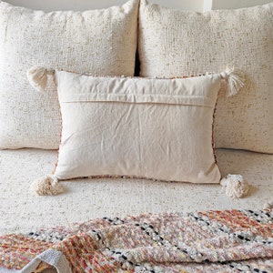 May include: A white throw pillow with tassels on the corners. The pillow is on a bed with a white textured bedspread and a multi-coloured throw blanket.